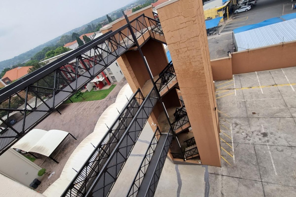 Aerial view of a commercial property featuring a metal pedestrian bridge connecting to a stairwell tower, overlooking an asphalt parking area with carports.