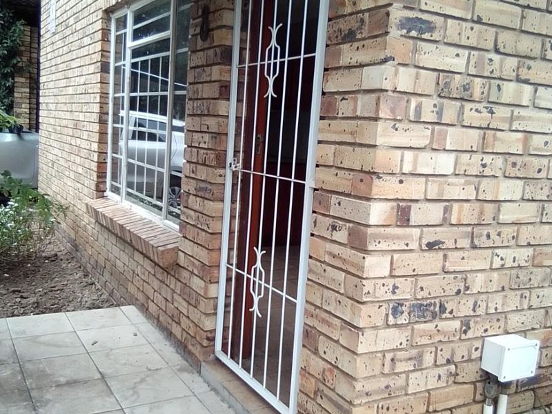 Front entrance of a face brick townhouse featuring a security gate, tiled porch area, and aluminium window frames.