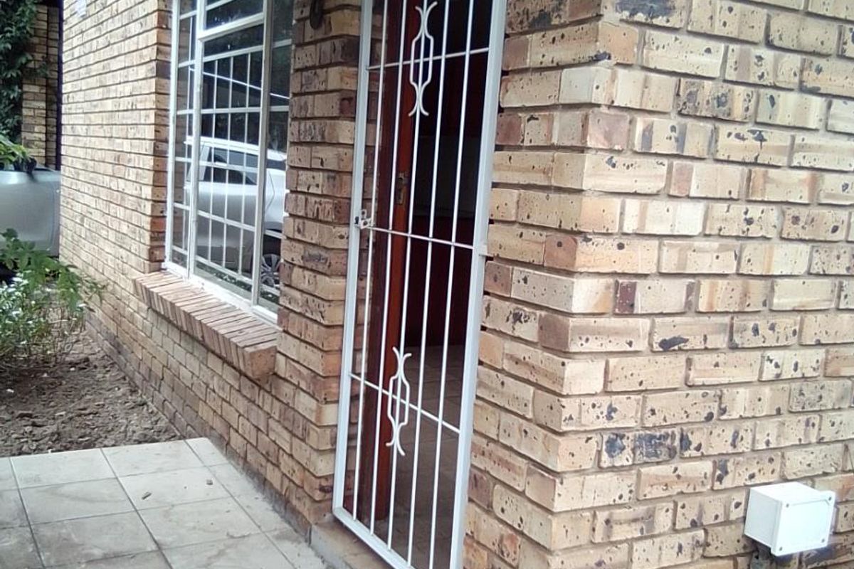 Front entrance of a face brick townhouse featuring a security gate, tiled porch area, and aluminium window frames.
