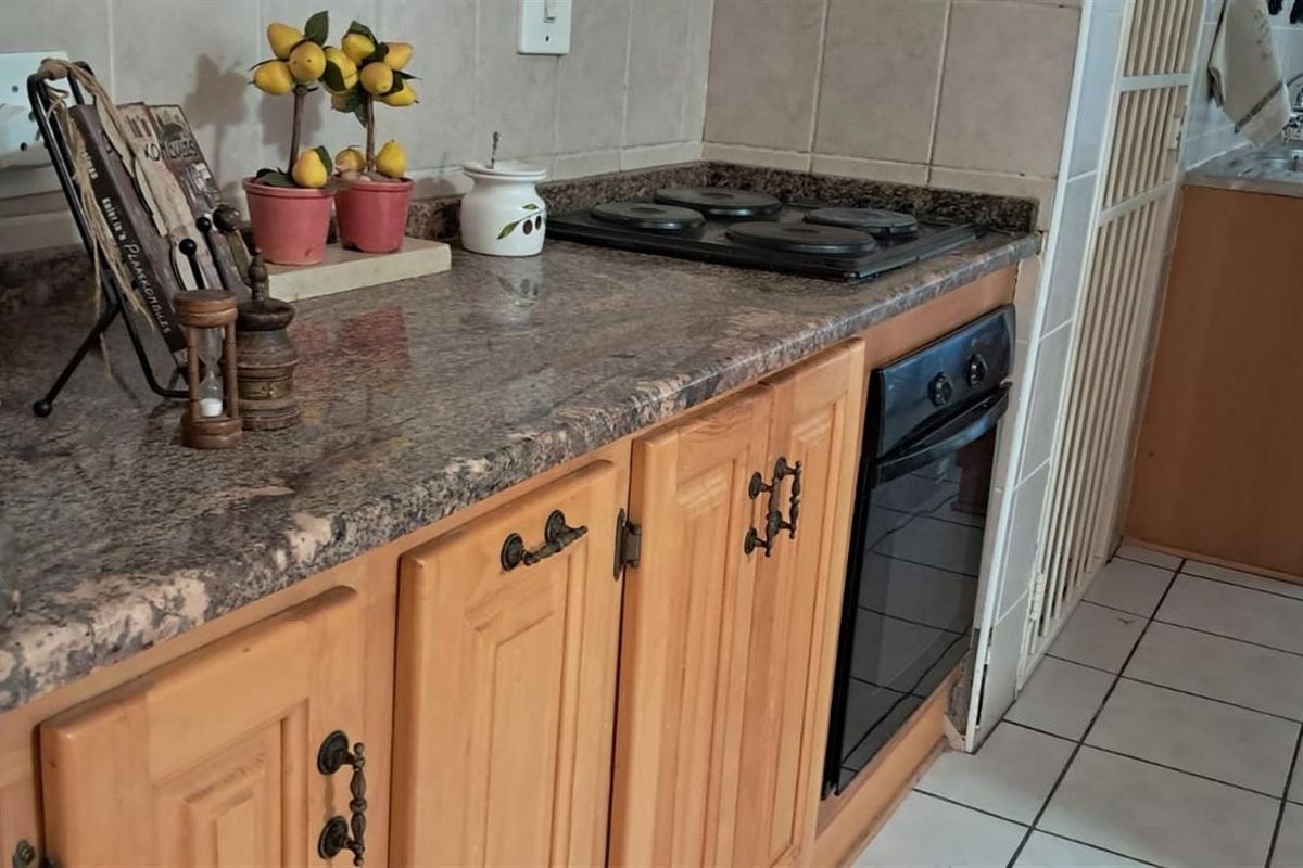 A functional kitchen area featuring granite countertops, built-in electric hob and oven, and wooden cabinetry with tiled flooring.