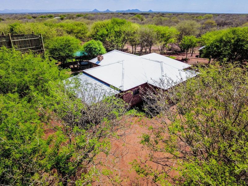 Aerial view of a farm property featuring a brick dwelling with a metal roof, surrounded by natural bushveld and an elevated wooden viewing deck.