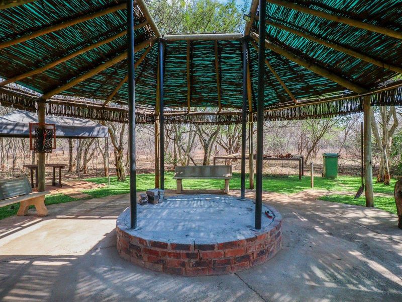 An outdoor entertainment area featuring a circular brick fire pit under a thatched roof structure with concrete flooring.