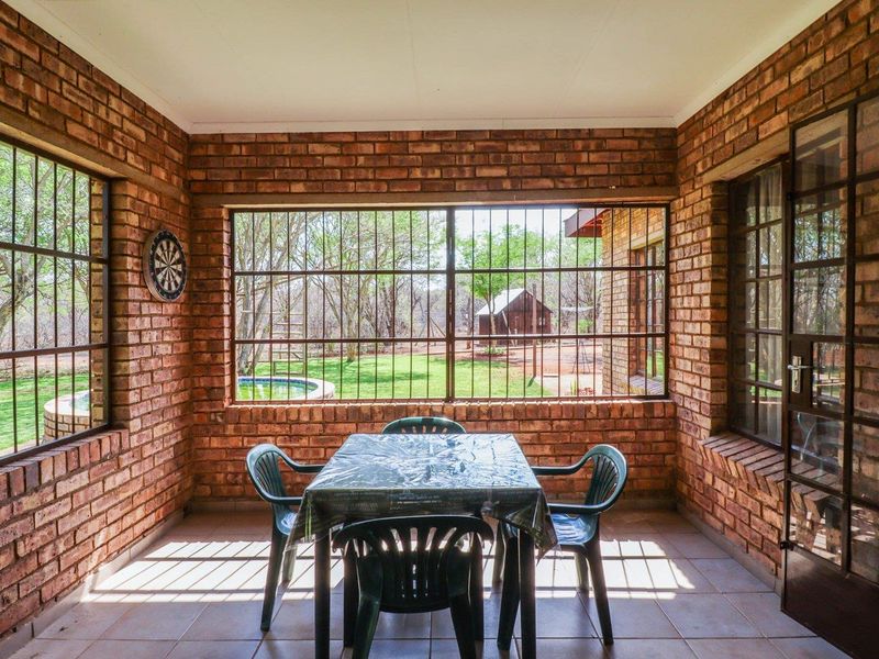 A covered patio area featuring exposed brick walls, tiled flooring, and large windows with security bars, offering views of the garden.