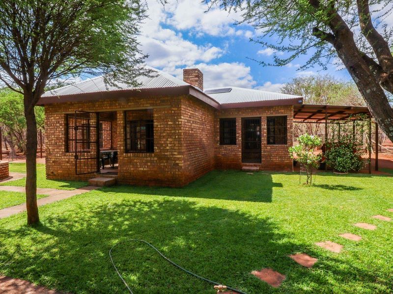Rear view of a face brick farmhouse featuring a corrugated metal roof, chimney, and a covered patio area set within a landscaped garden.