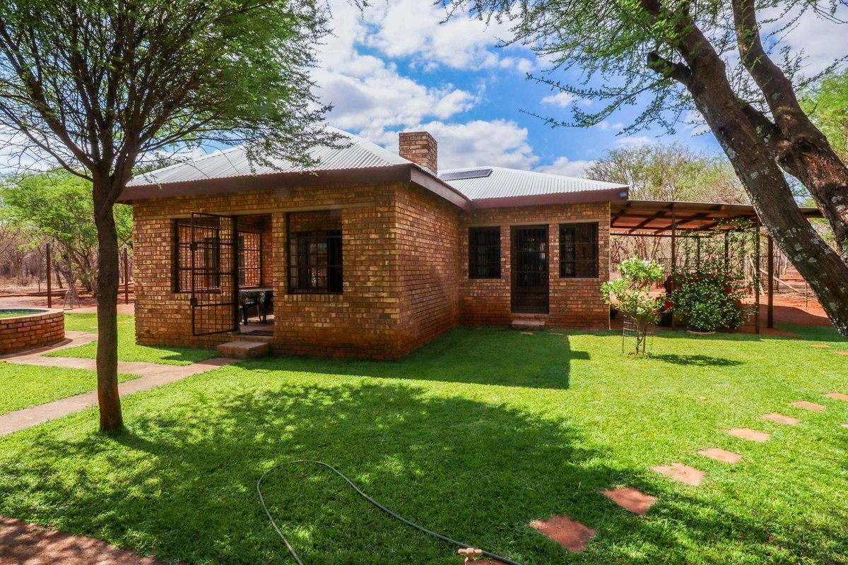 Rear view of a face brick farmhouse featuring a corrugated metal roof, chimney, and a covered patio area set within a landscaped garden.