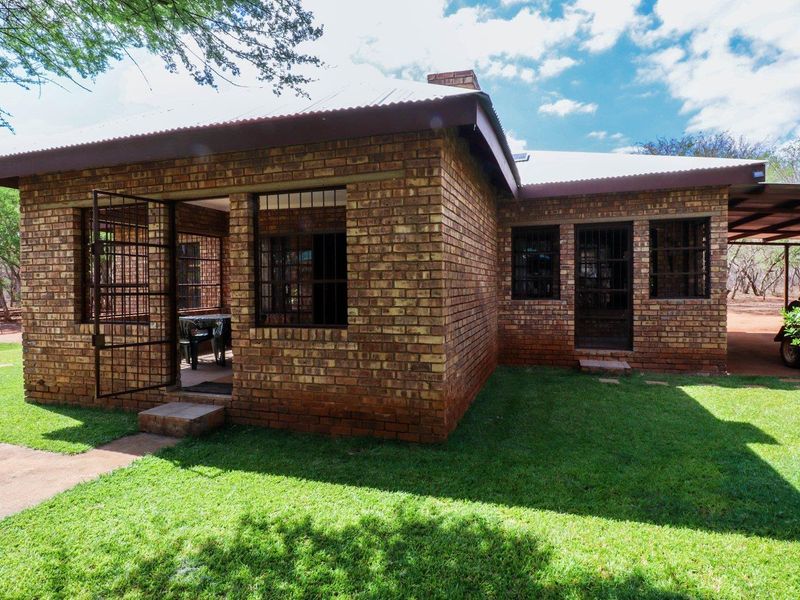 A rear view of a face brick farm cottage featuring a covered patio, security gates, and an attached carport set within a garden.