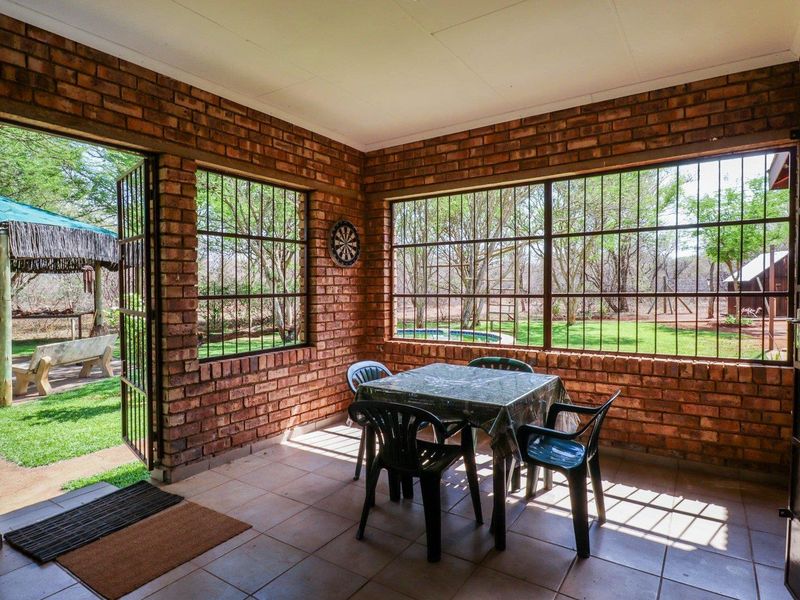 A covered patio area featuring exposed brick walls, tiled flooring, and large windows with security bars, offering views of the garden and pool area.