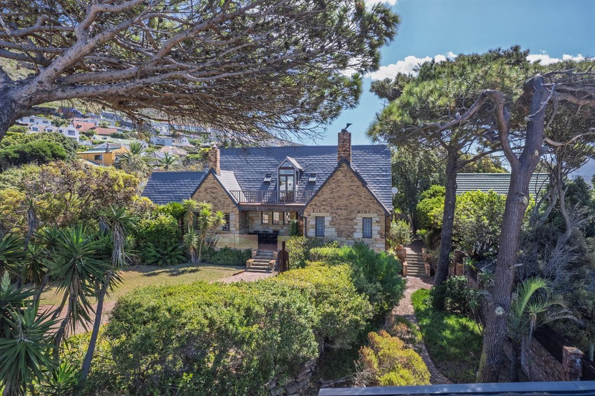 A charming stone-clad house featuring a pitched slate roof, dormer windows, and a balcony, set within a lush, mature garden with established trees.
