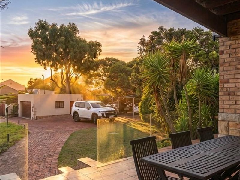 A covered patio featuring tiled flooring and glass balustrades, overlooking a landscaped garden and driveway at sunset.