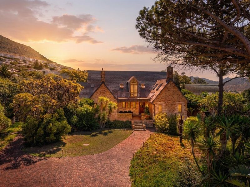 A charming stone-clad residence featuring a pitched slate roof, balcony, and established garden, set against a scenic mountain backdrop at sunset.