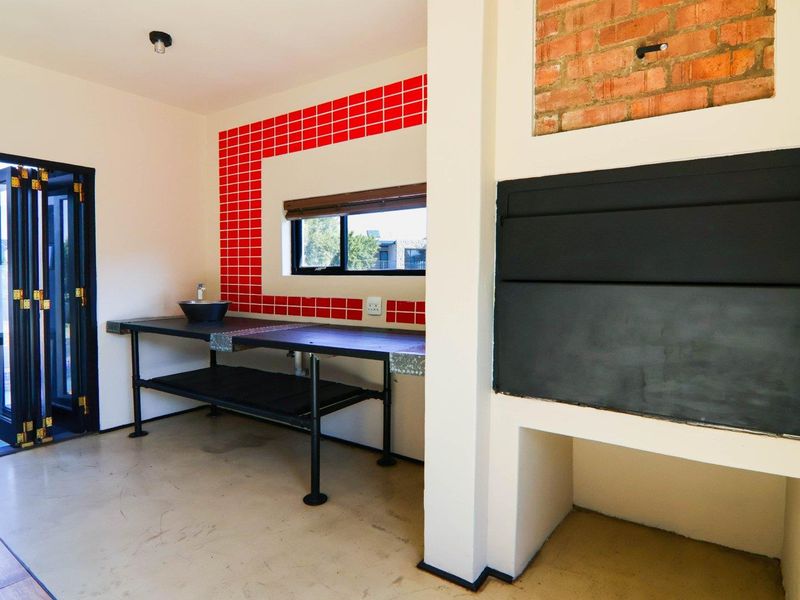 Covered patio area featuring a built-in braai, industrial-style metal prep counter with basin, red tiled splashback, and bi-fold doors leading to the pool area.