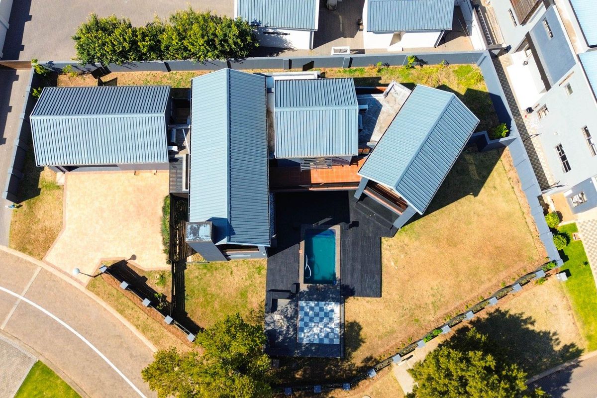 Aerial view of a modern residential property featuring a metal roof, swimming pool, timber decking, and a paved driveway within a secure estate.