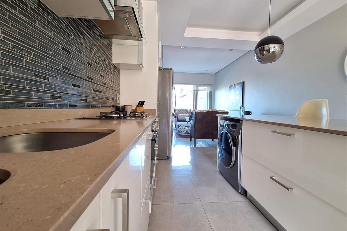 Modern galley-style kitchen featuring sleek white cabinetry, stone countertops, a mosaic tile backsplash, and integrated laundry appliances.