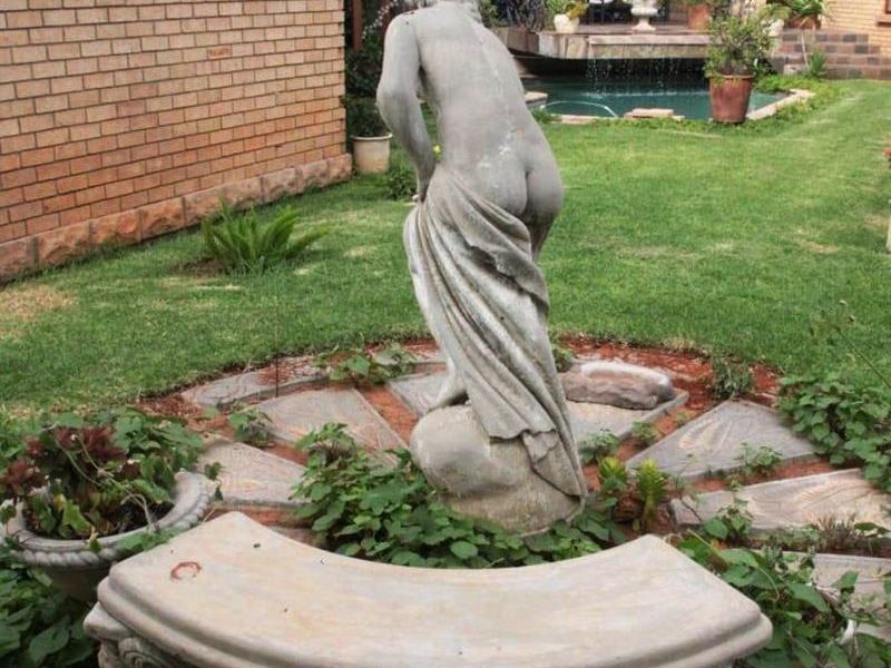 A landscaped garden area featuring a stone bench and decorative statue, with a view towards the rear patio and swimming pool.
