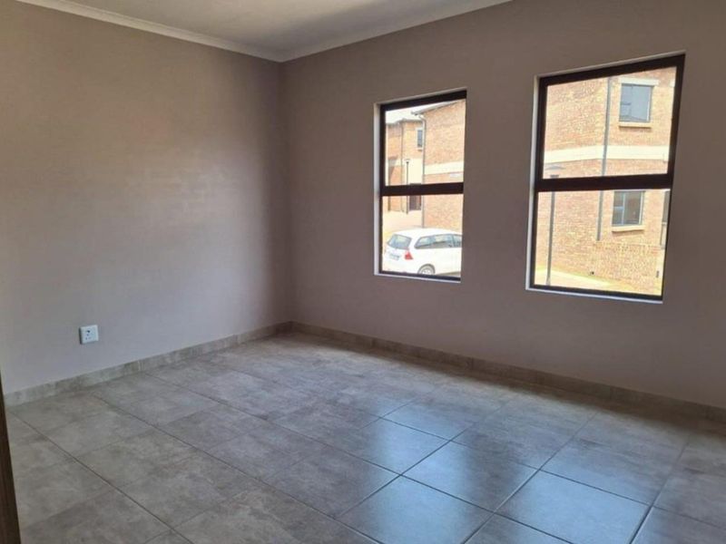 A bright, empty bedroom featuring neutral wall tones, modern tiled flooring, and large aluminium-framed windows providing natural light.
