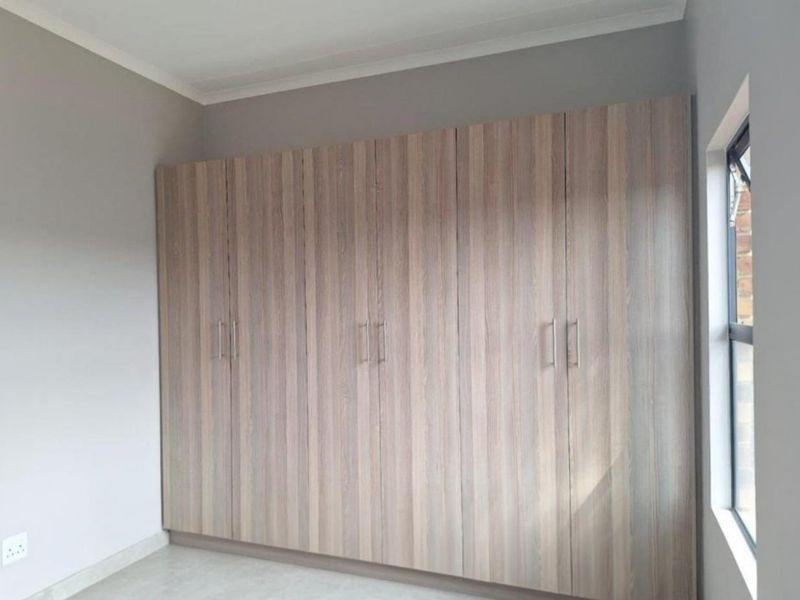 A clean, empty bedroom featuring modern wood-finish built-in cupboards, neutral wall paint, and a side window providing natural light.
