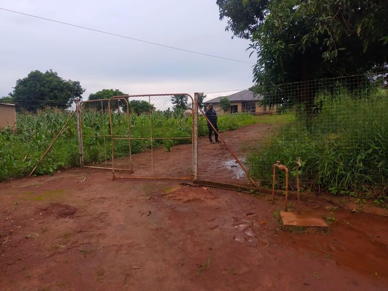 Front view of a residential plot featuring a metal gate, wire fencing, and an outdoor water tap on a dirt driveway.