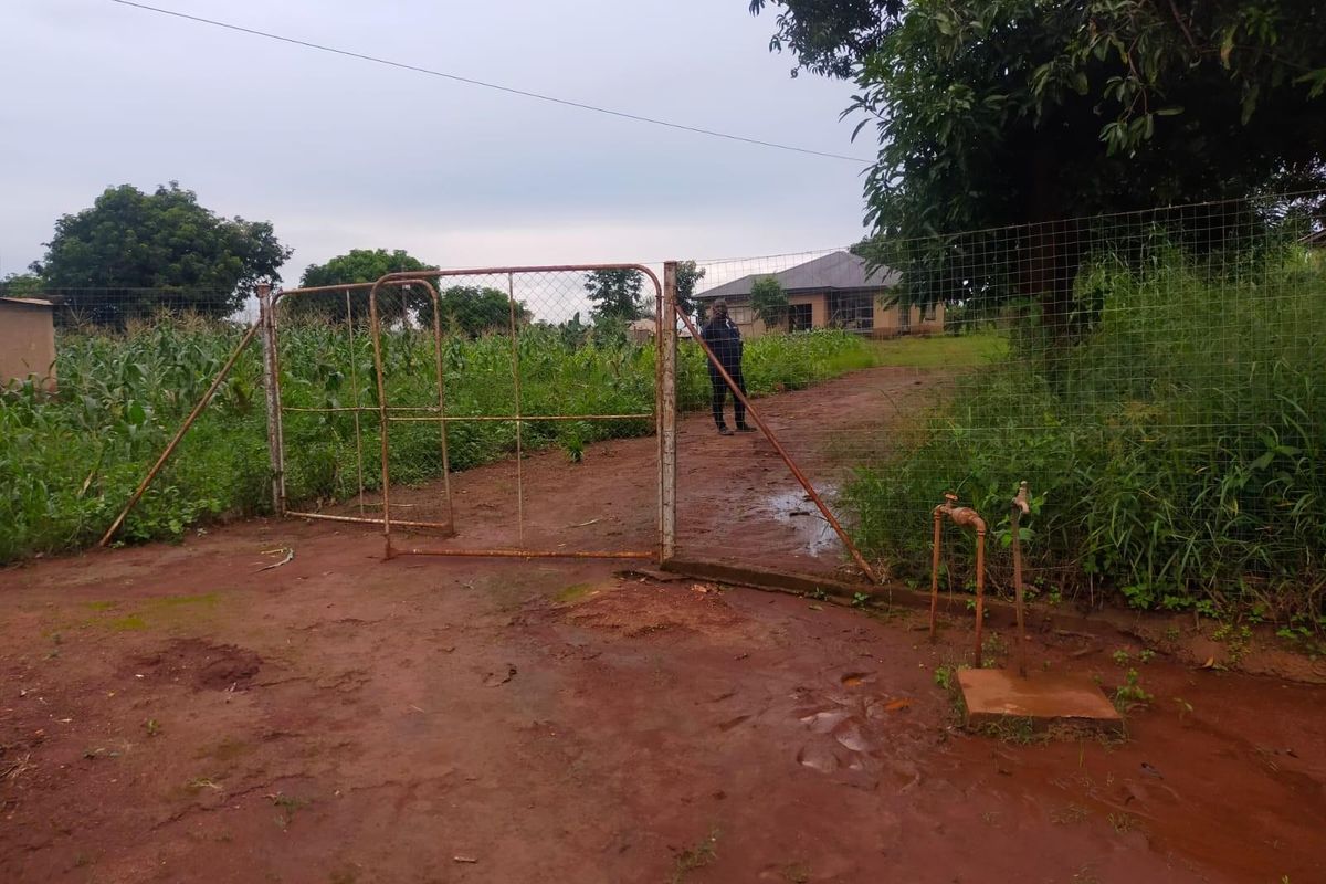 Front view of a residential plot featuring a metal gate, wire fencing, and an outdoor water tap on a dirt driveway.