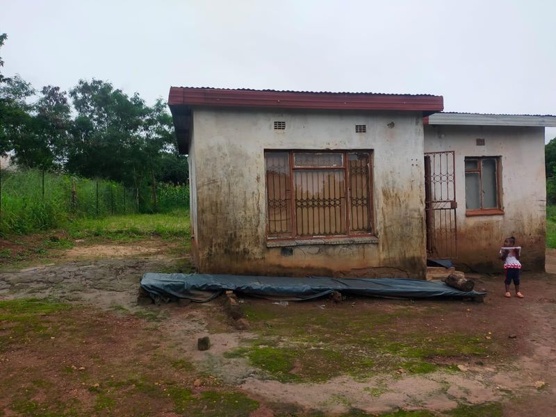 Front view of a residential structure showing significant exterior wall weathering and deterioration, featuring metal security bars and a corrugated roof.