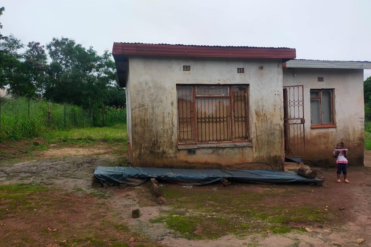 Front view of a residential structure showing significant exterior wall weathering and deterioration, featuring metal security bars and a corrugated roof.