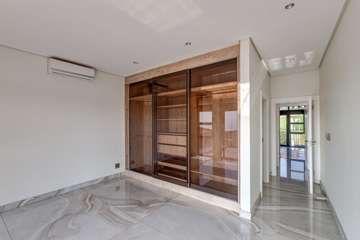 A modern bedroom featuring large format marble-look floor tiles, recessed ceiling lighting, a wall-mounted air conditioning unit, and a built-in floor-to-ceiling glass-fronted wardrobe.