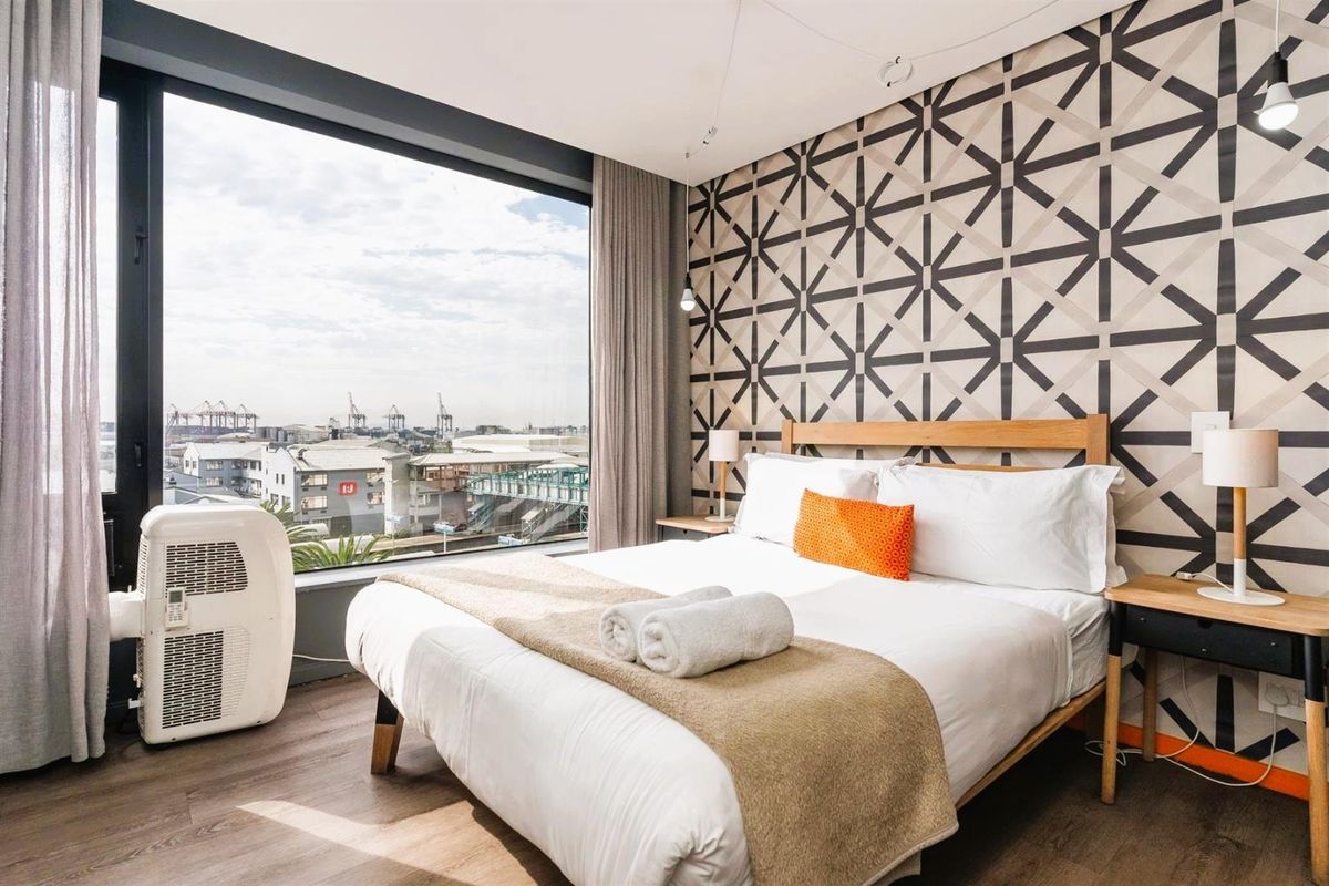 A modern bedroom featuring floor-to-ceiling windows with urban views, patterned accent wall, and wood-look flooring.