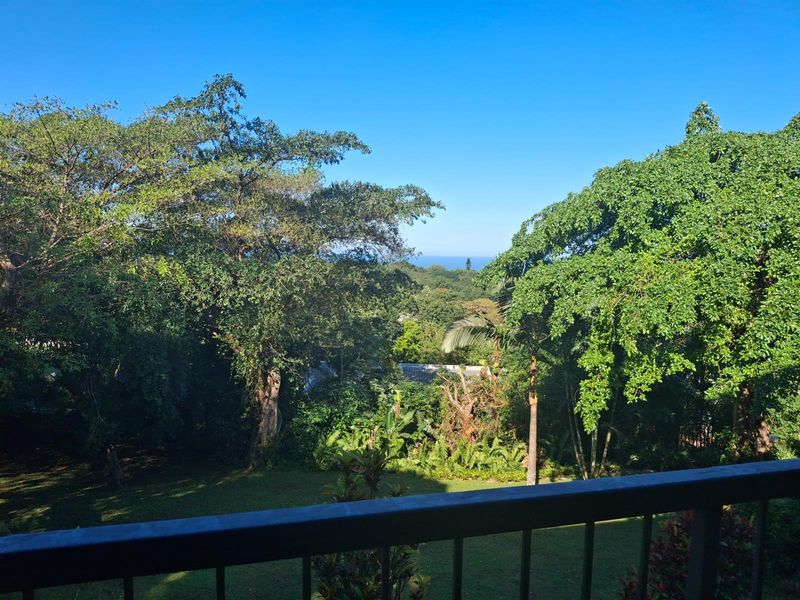 A scenic view overlooking a lush green garden and mature trees, with a distant glimpse of the ocean horizon.
