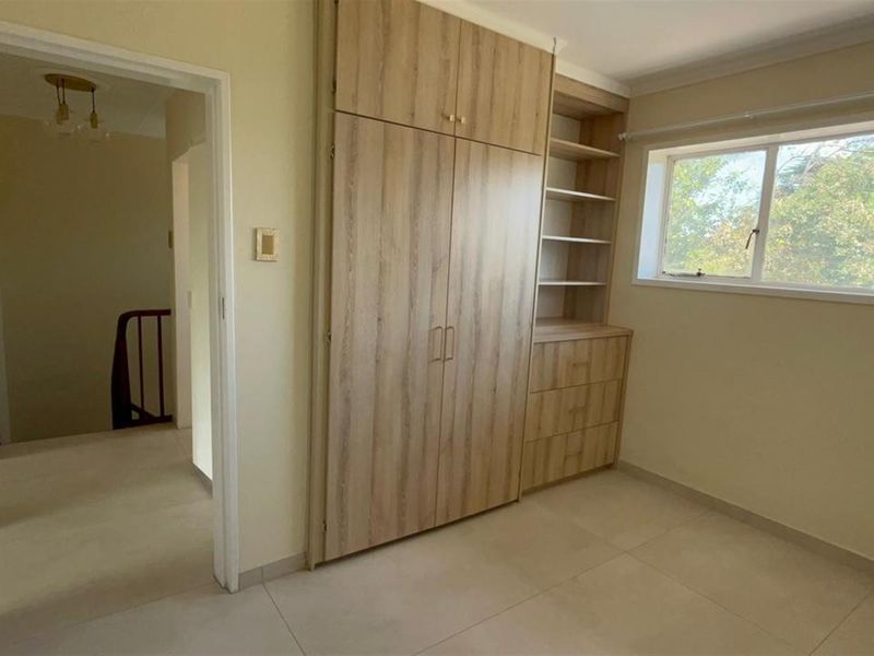 A bright, empty bedroom featuring modern light wood built-in cupboards with integrated shelving and neutral tiled flooring.