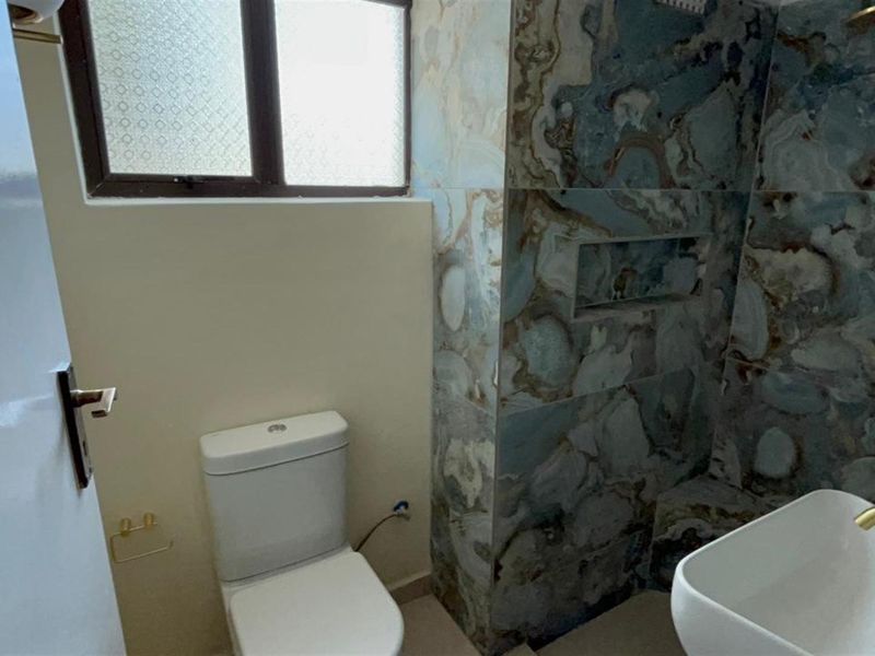 A compact bathroom featuring a modern basin, toilet, and a shower enclosure with decorative marble-effect tiling and brass fixtures.