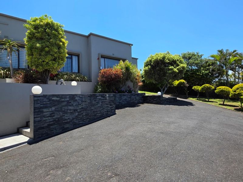 A well-maintained front exterior featuring a paved driveway, stone-clad retaining wall, and established garden landscaping under a clear blue sky.