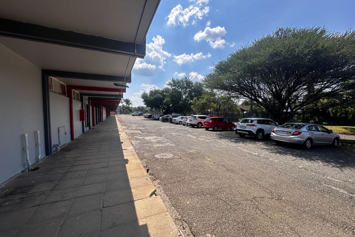 Exterior view of a commercial property featuring a paved walkway alongside a large asphalt parking area with mature trees.