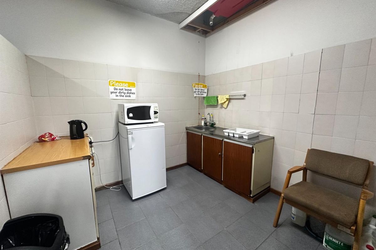 A functional commercial kitchenette featuring a stainless steel sink, tiled splashbacks, and basic cabinetry, currently showing signs of wear and an exposed ceiling area.