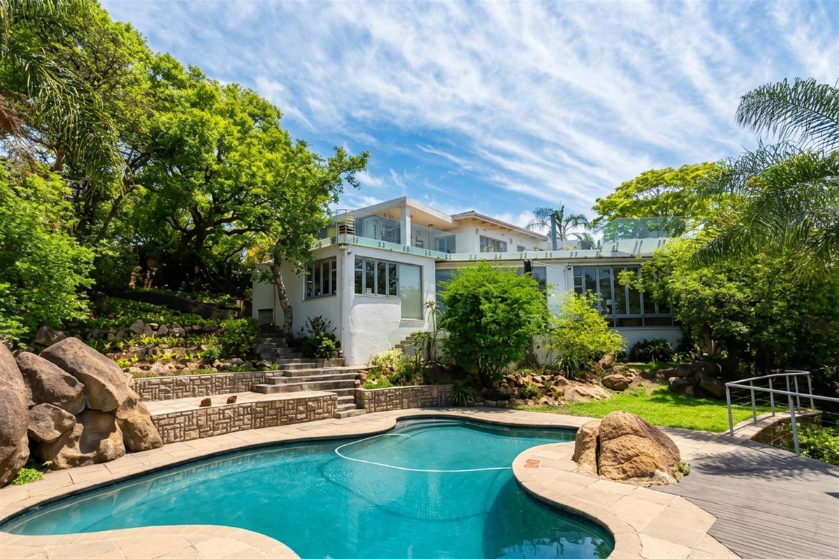 Rear exterior view featuring a swimming pool, stone patio, landscaped garden, and a multi-level home with glass balustrades.