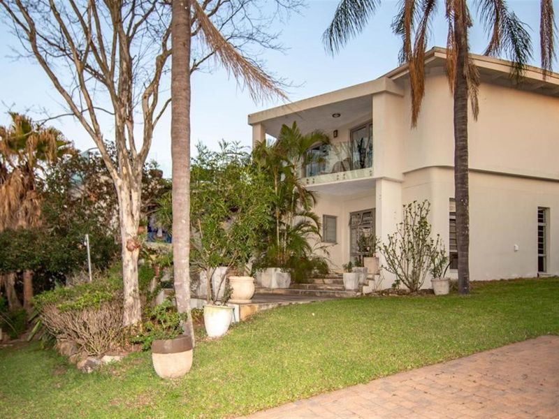 A two-story residential property featuring a paved driveway, landscaped garden, and an upper-level balcony with a glass balustrade.