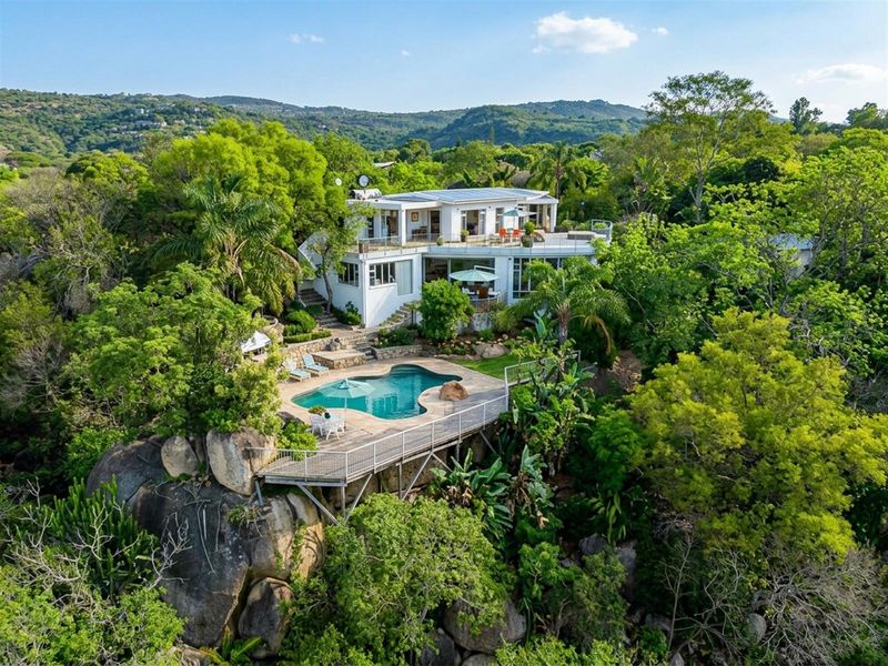 Aerial view of a multi-level residence featuring a swimming pool, raised deck, and solar panels, set within a lush, elevated landscape.