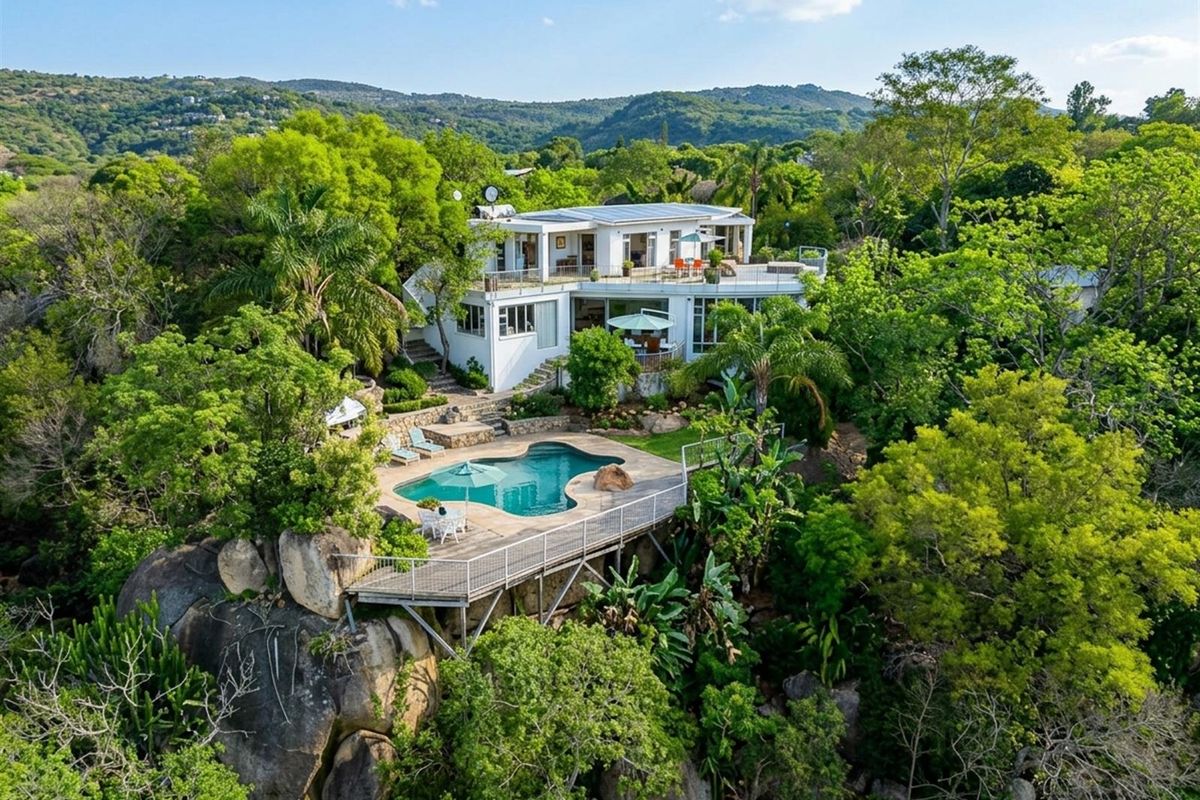 Aerial view of a multi-level residence featuring a swimming pool, raised deck, and solar panels, set within a lush, elevated landscape.