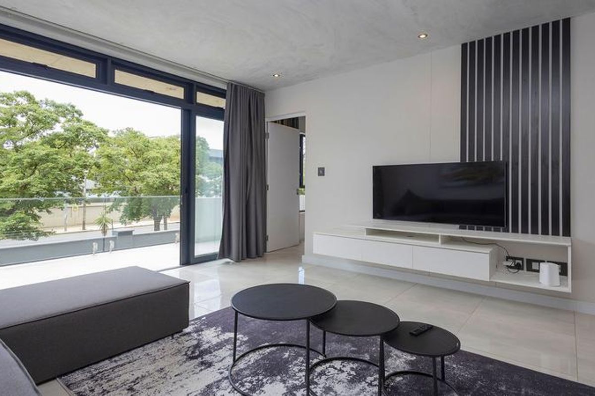 Modern living room featuring sleek tiled floors, recessed lighting, a wall-mounted media console, and large sliding doors opening to a balcony with a glass balustrade.