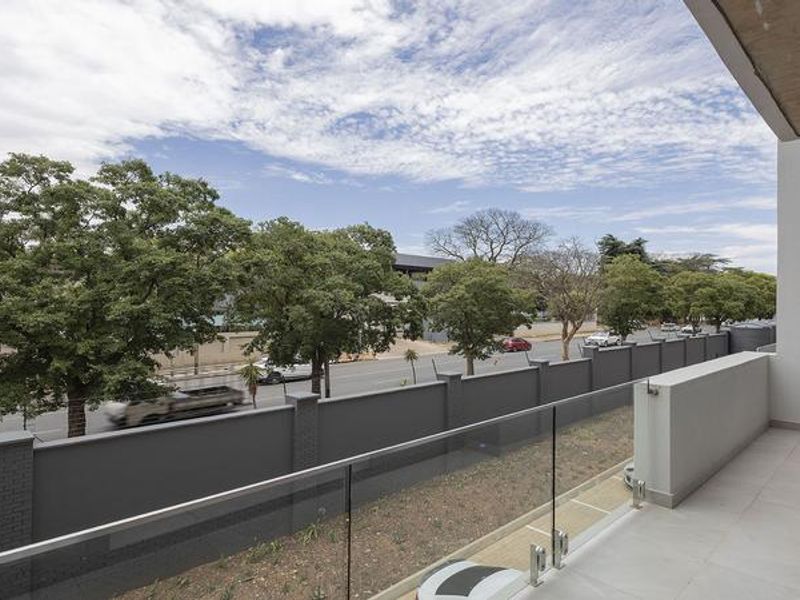 A modern, covered balcony featuring a sleek glass balustrade, tiled flooring, and recessed ceiling lighting, offering an elevated view of the street and surrounding trees.