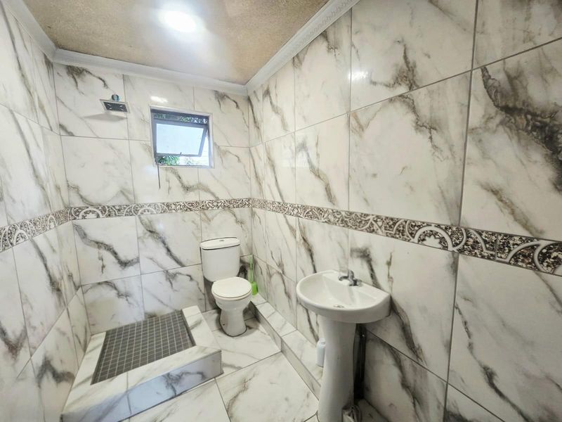 A compact bathroom featuring marble-look wall tiles, a pedestal basin, a toilet, and a tiled shower area with a fixed shower head.