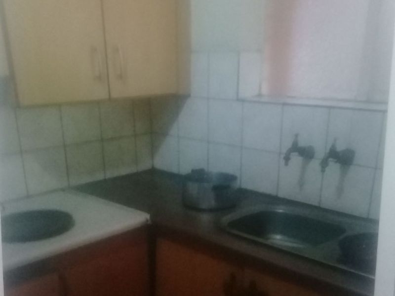 A compact kitchen area featuring dated built-in cabinetry, a stainless steel sink with wall-mounted taps, and tiled splashbacks.