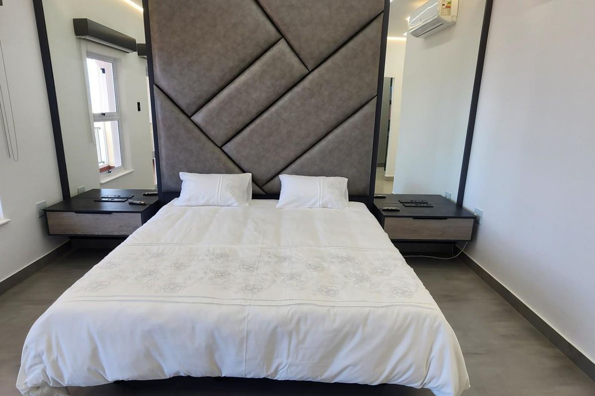 Modern bedroom featuring a large upholstered headboard, integrated bedside cabinetry, and a wall-mounted air conditioning unit.
