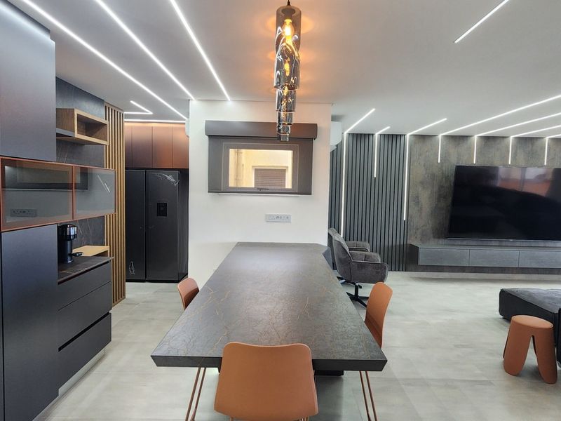Modern open-plan living area featuring a sleek kitchen with dark cabinetry, integrated appliances, linear LED ceiling lighting, and a contemporary media wall.