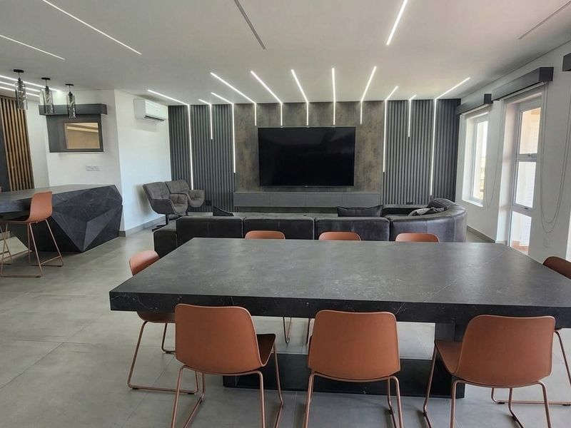 Modern open-plan living area featuring a sleek kitchen with breakfast bar, dining space, and integrated linear LED ceiling lighting, opening onto a private balcony.