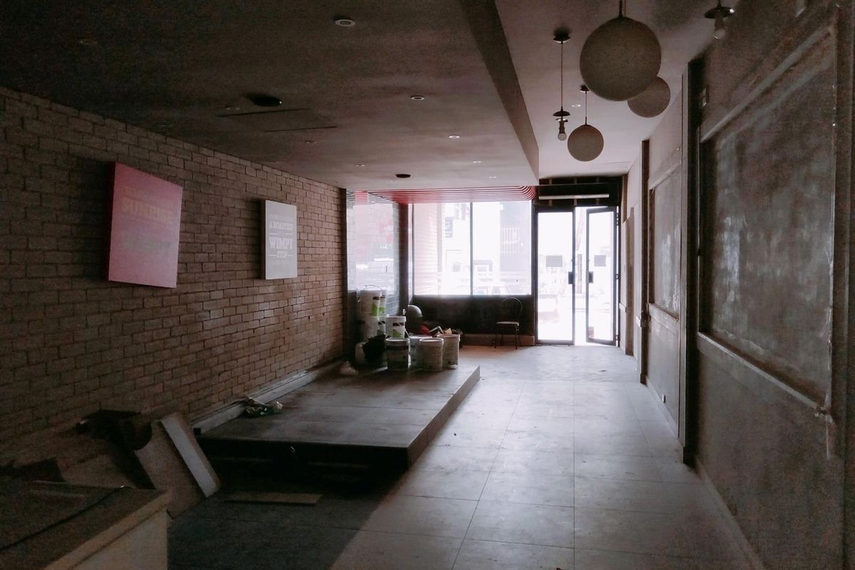 An open-plan commercial space featuring exposed brick walls, tiled flooring, a raised platform, and modern pendant lighting, currently in a vacant, unfinished state.