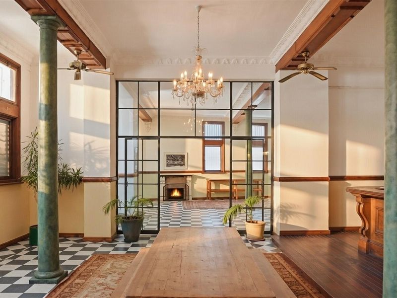 Spacious commercial interior featuring decorative columns, a classic chandelier, checkerboard flooring, and a large glass-paneled partition leading to a secondary room with a fireplace.