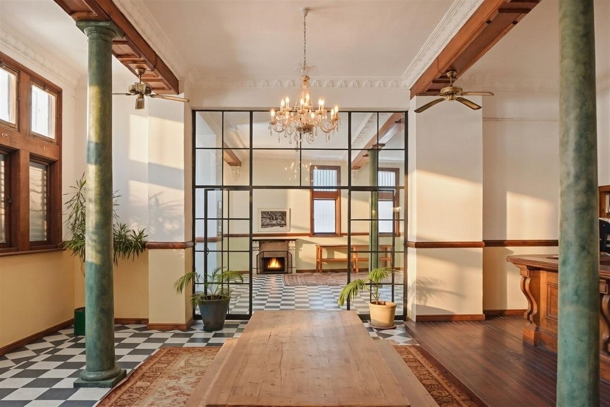 Spacious commercial interior featuring decorative columns, a classic chandelier, checkerboard flooring, and a large glass-paneled partition leading to a secondary room with a fireplace.