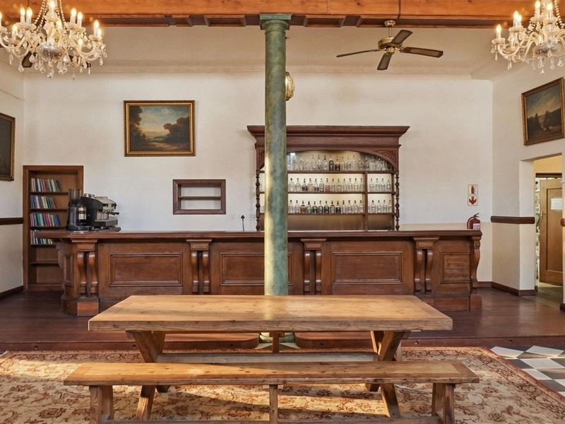 A classic commercial bar area featuring a substantial wooden bar counter, built-in shelving, decorative chandeliers, and a central structural column.