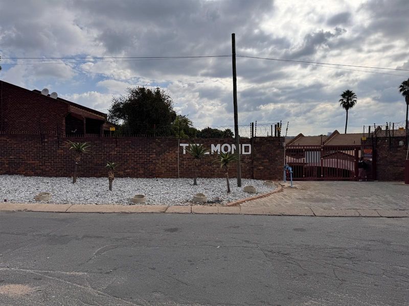 Street view of the apartment complex entrance featuring a brick boundary wall, electric fencing, and a secure gated driveway.