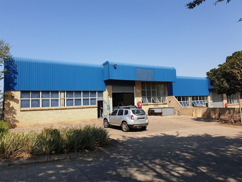 Industrial warehouse facility featuring a blue corrugated metal roof, brick facade, large windows, and a paved forecourt area.