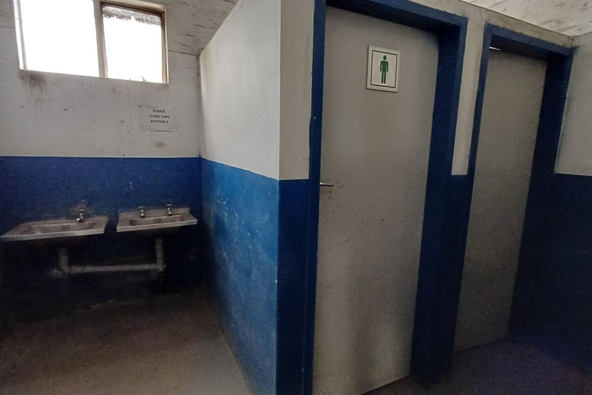 Industrial restroom facility featuring dual stainless steel basins and partitioned cubicle doors, showing significant signs of wear and dated finishes.